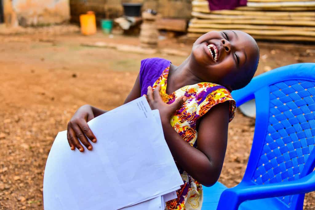 Girl wearing a purple and orange patterned dress. She is sitting in a blue plastic chair outside her home and is holding a letter from her sponsor.