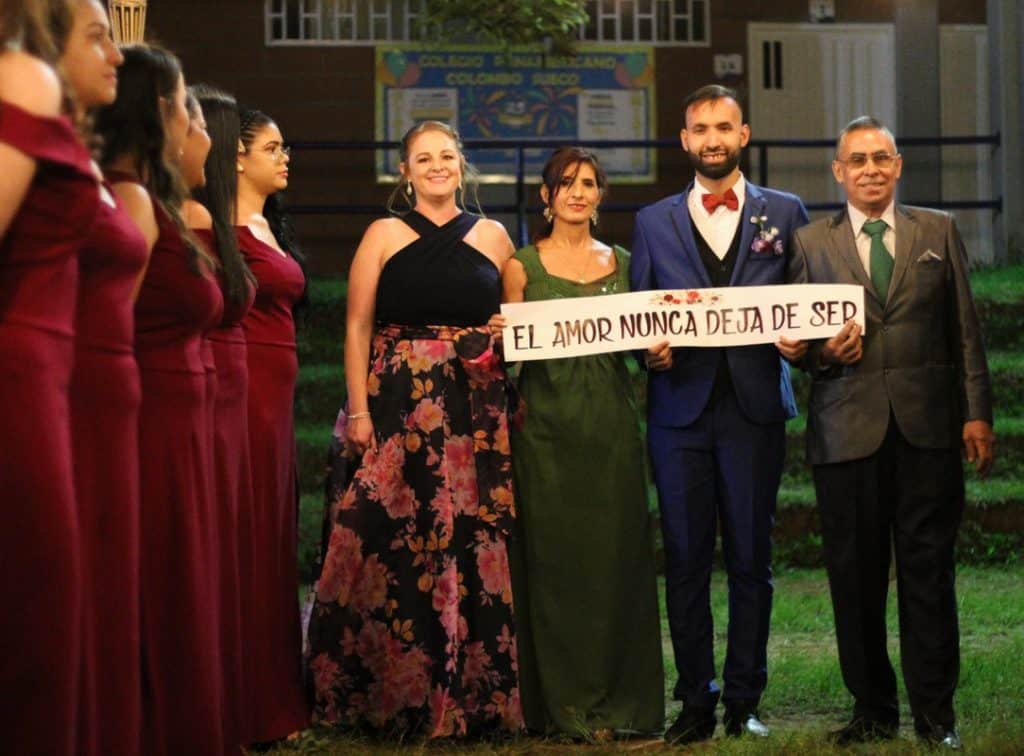 Two men and two women stands next to each other outdoors at a wedding in Colombia. They are holding a sign that says "El amor nunca deja de ser," which translates to "Love never ceases to be" in English. There are bridesmaids wearing red dresses lined up near them.