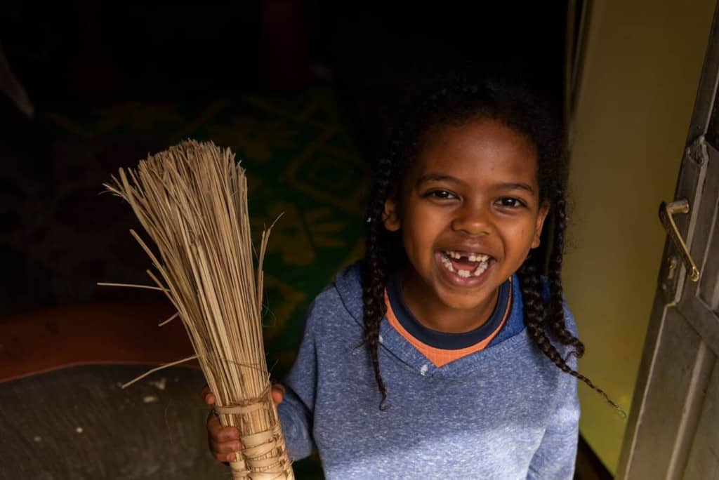 Tensae is wearing a blue sweater. She is holding a broom and is standing inside her home.