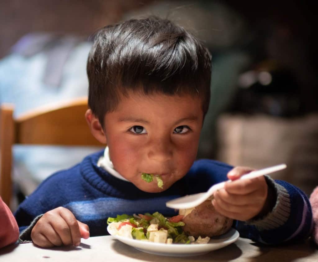 Yomel is wearing a blue sweater. He is sitting at a table inside his home and is eating a salad and potatoes.