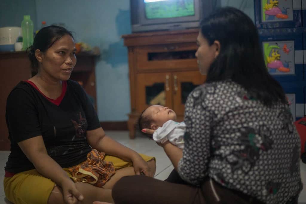 Woman in a black shirt, is sitting at home on the floor with a project staff member who is conducting a home visit after the flood had receded. The staff member is holding baby Gabrian, who is sleeping.