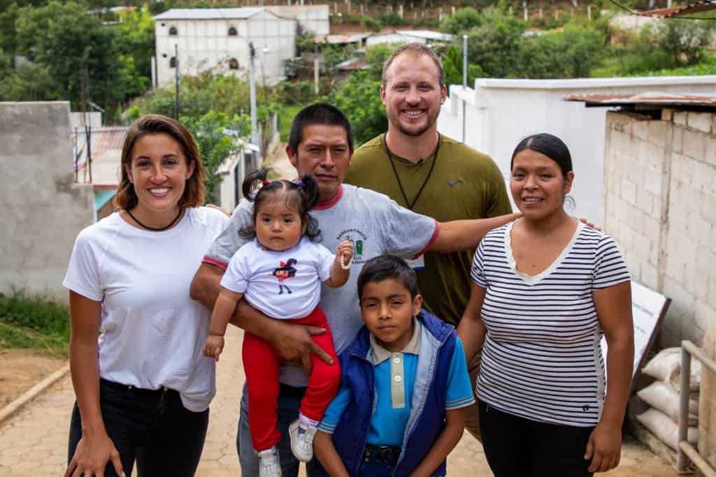 Nate (in khaki green) and Lexi (in white) are standing on a set of steps posing for a photo with a family. The family consists of a dad, mom and two children, a baby girl in red and white, and a boy in blue. There are white buildings on either side of them. 
