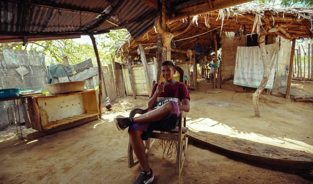 A youth, boy, sits in a wooden chair giving a thumbs up. The floor beneath him is sand, and you can see the thatch roof of the home where he lives in Colombia. 