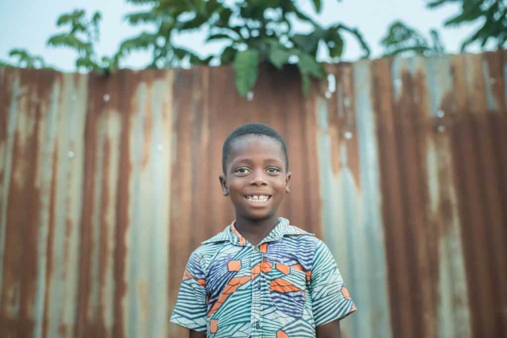 Boy wearing a blue and orange pattern shirt smiles standing outside with a metal rusted fence in the background.