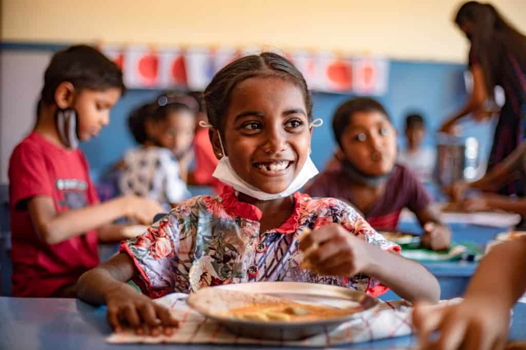 Theresa is wearing a colorfully patterned dress. She is sitting down at a table with a plate of food in front of her. The meal for the day included dosai, lentils curry and coconut chutney, most of the children’s favorite dish.