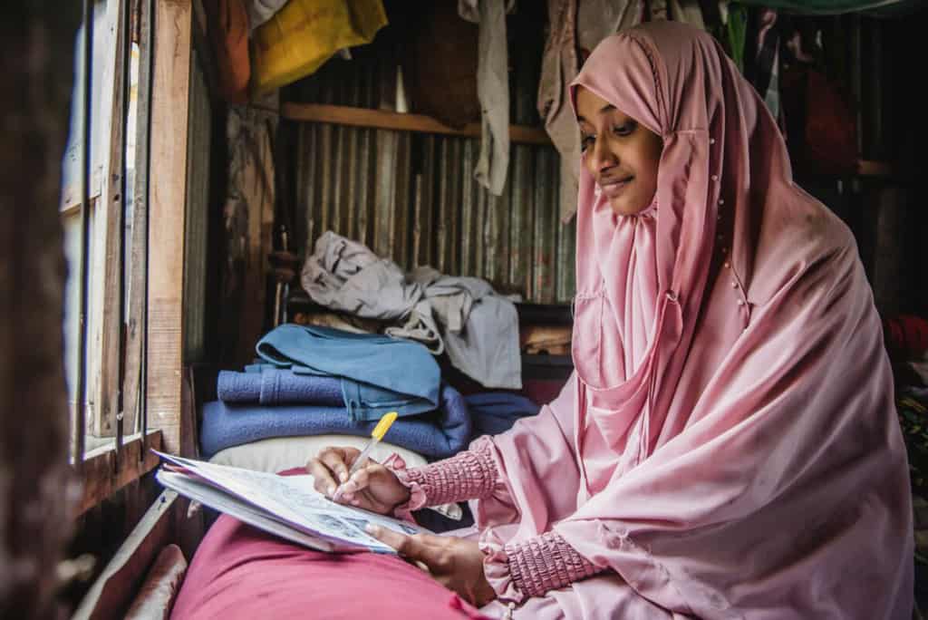 Woman wearing a pink dress and head covering. She is sitting in front of a window in her home and is writing a letter to her sponsor.