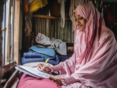 Woman wearing a pink dress and head covering. She is sitting in front of a window in her home and is writing a letter to her sponsor.