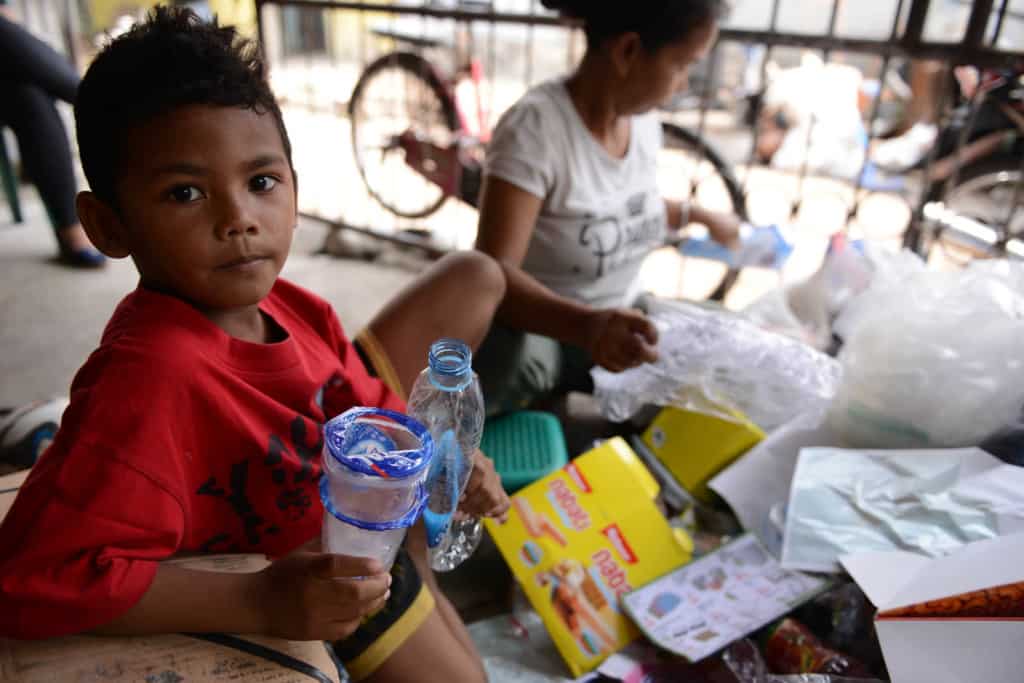7-year-old boy in Jakarta holds empty plastic cups and a plastic bottle and makes eye contact. An adult female behind him sorts recyclables.
