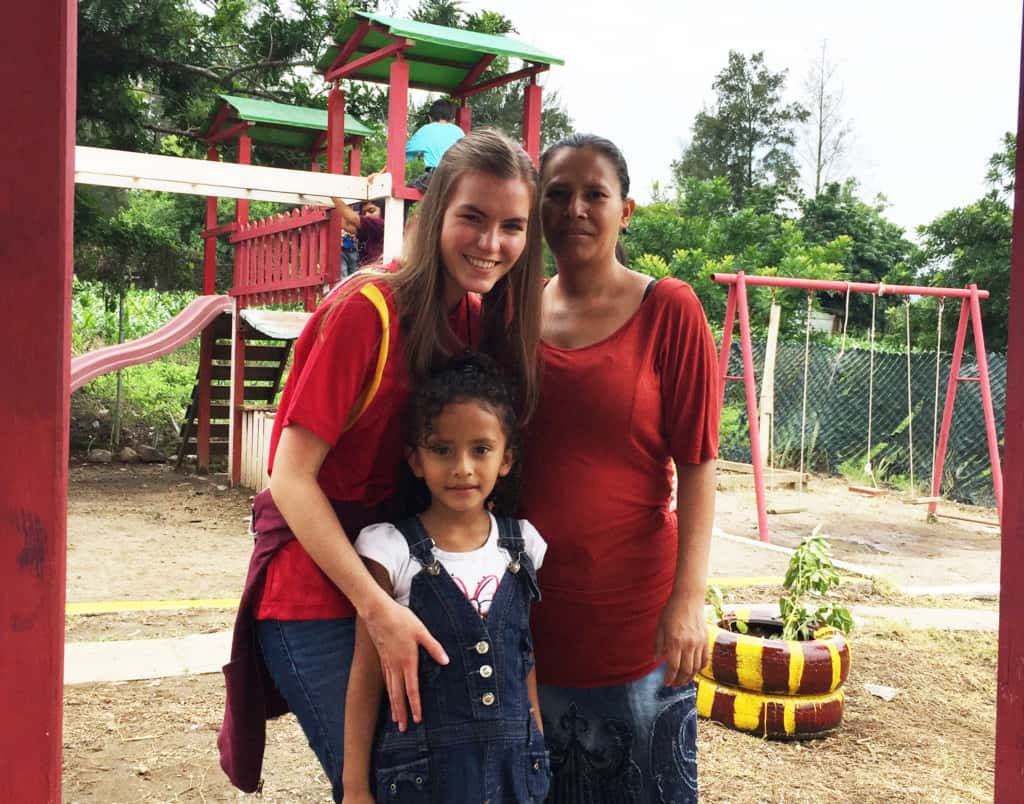Two women, Hayli and Imelda, stand with a young girl, Genesis. There is a swing set in the background. The women are wearing red shirts, and the girl is wearing overalls.