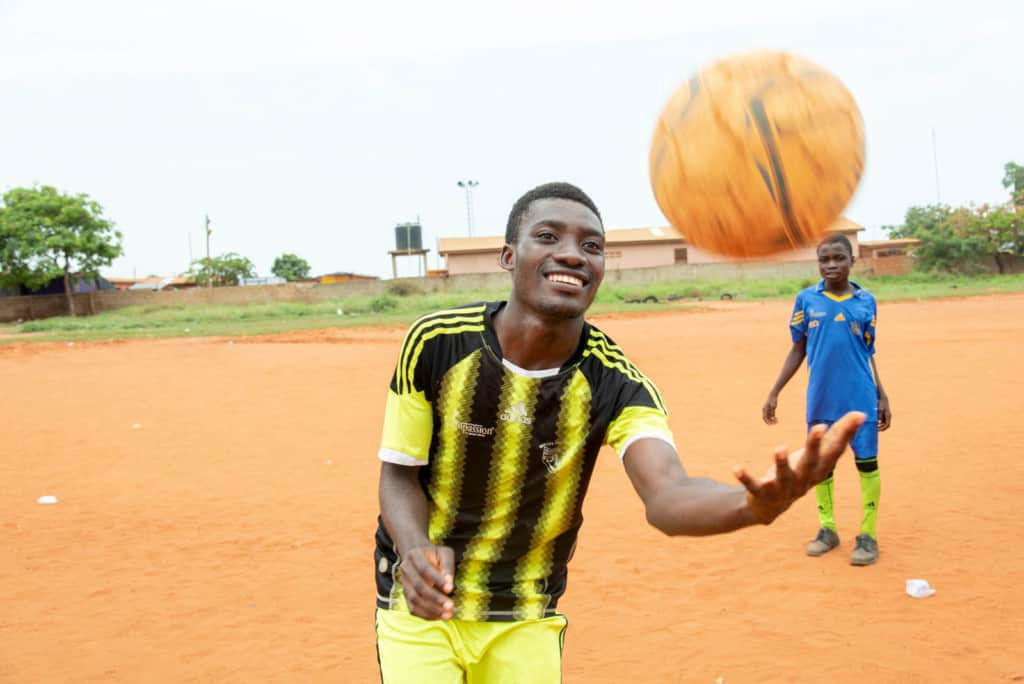 An action shot of a twenty-one-year-old adult male wearing a black and yellow soccer uniform, jersey, juggling, throwing a soccer ball in the air. He is standing on a dirt soccer field. 