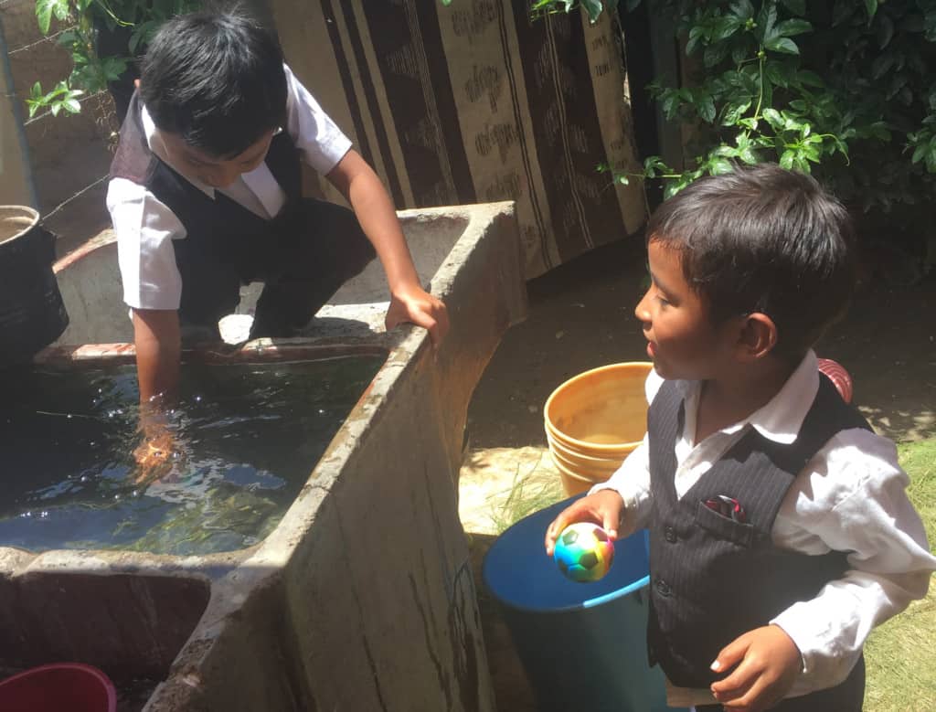 Two young boys are wearing vests and white collared shirts. One is holding a toy soccer ball. One is putting his hand into an outdoor sink of water.