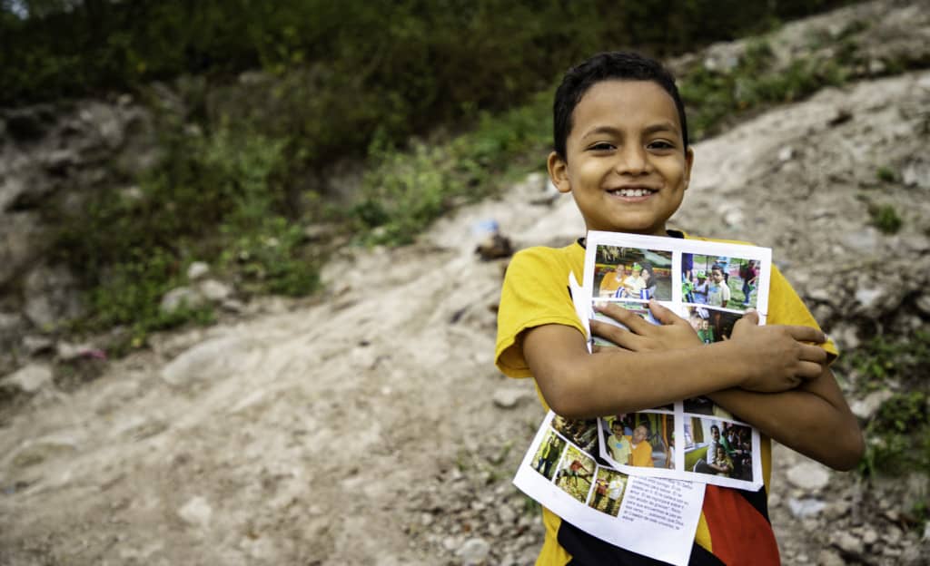 Jairo is wearing a yellow shirt and jeans. He is standing outside his home and is holding his sponsor letters close to his chest.