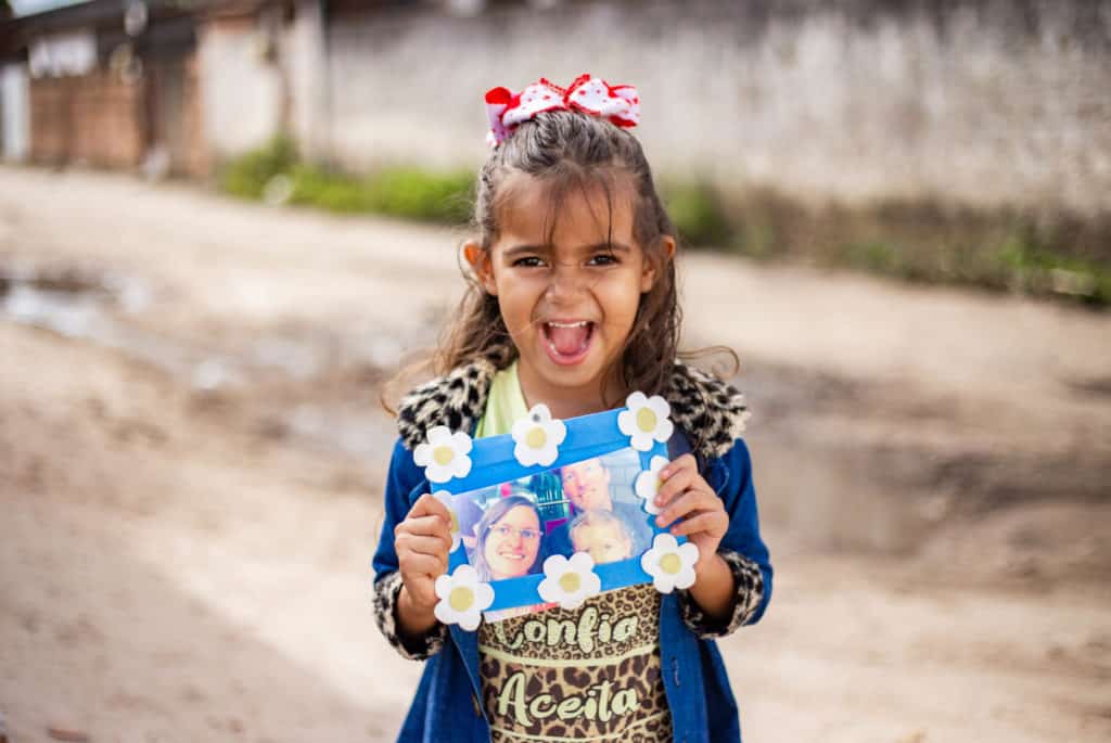 Débora is wearing a yellow shirt with a black pattern on it and a blue jacket. She is standing outside her home and is holding up a picture of her sponsors.