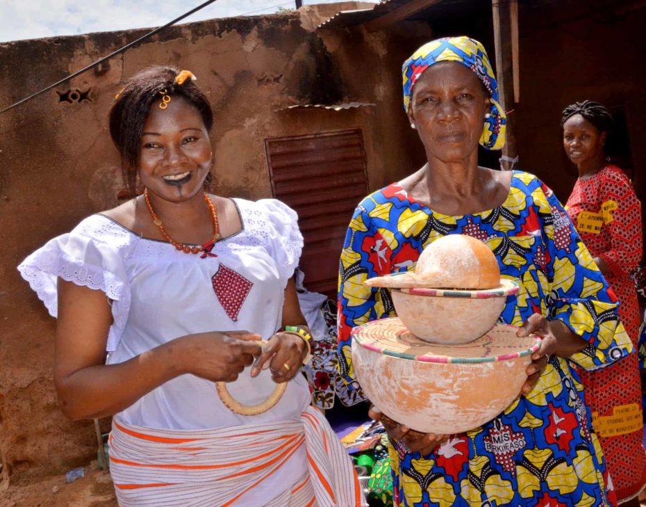 Odette and another woman in Burkina Faso are wearing brightly colored clothing and standing outdoors together. One woman is holding two ceramic bowls