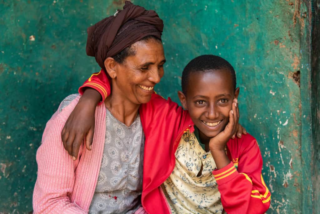 Abel, in a red jacket, is at home sitting and posing for a picture with his mom, Astede, who is wearing a pink sweater. She is looking at him and smiling, and he has his arm around her shoulders. The wall behind them is green. Abel faced a cancer battle and won.