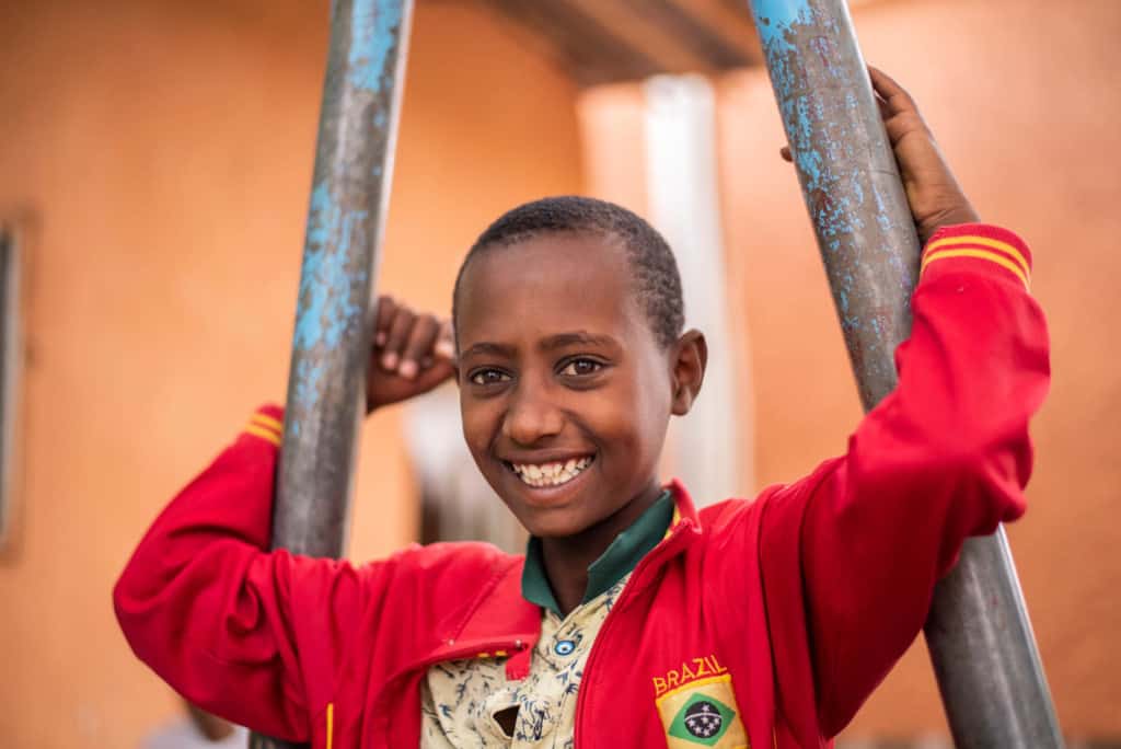 Abel, in a red jacket, is outside the church smiling and holding onto the poles of a piece of playground equipment. The building in the background is orange.