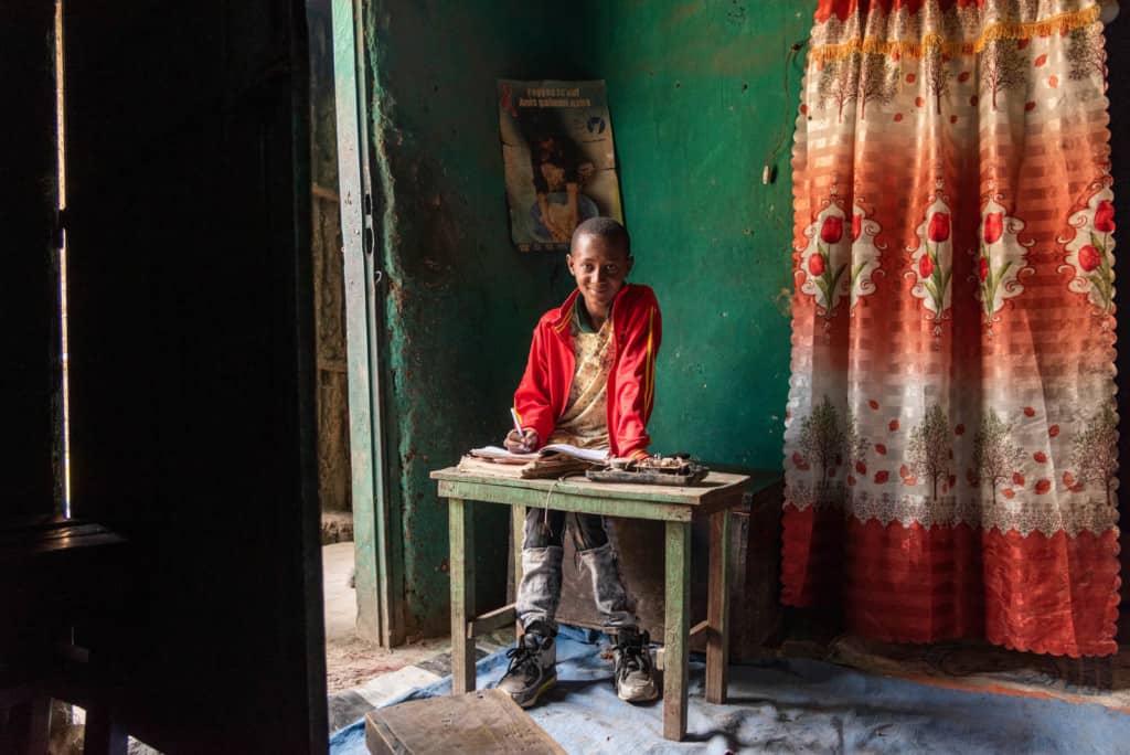 Abel, in a red jacket, is at home sitting at a small green wood desk. The walls behind him are green and there is an orange and red curtain hanging next to him. 