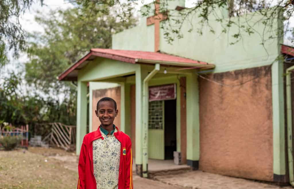 Abel, in a red jacket, is smiling and standing outside in front of a pale green church.