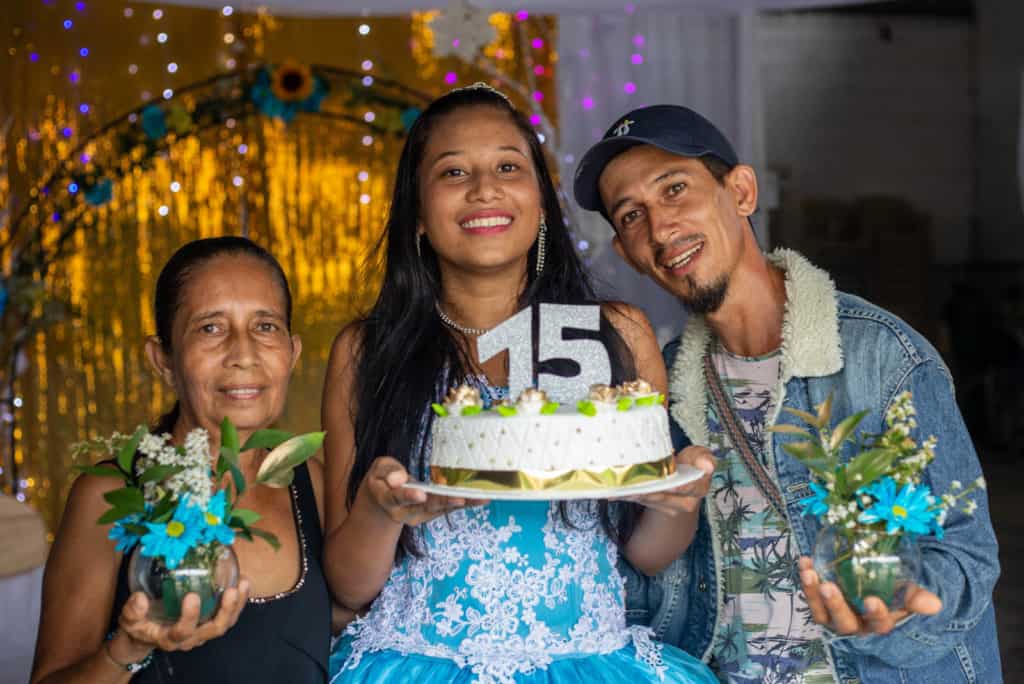 Girl wearing a blue dress and is holding a cake with a large 15 on it. She is standing with her father and grandmother in front of a gold curtain.