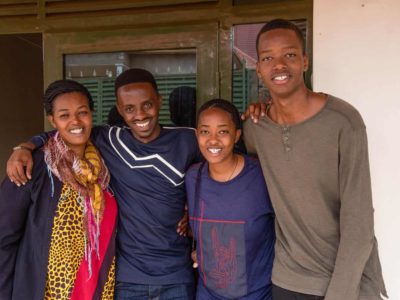 Christian is wearing a navy blue shirt with gray stripes down the center. He is outside his aunt's home with his Aunt Pascasie, and his cousins, Cynthia and Mugisha. They have their arms around each other.