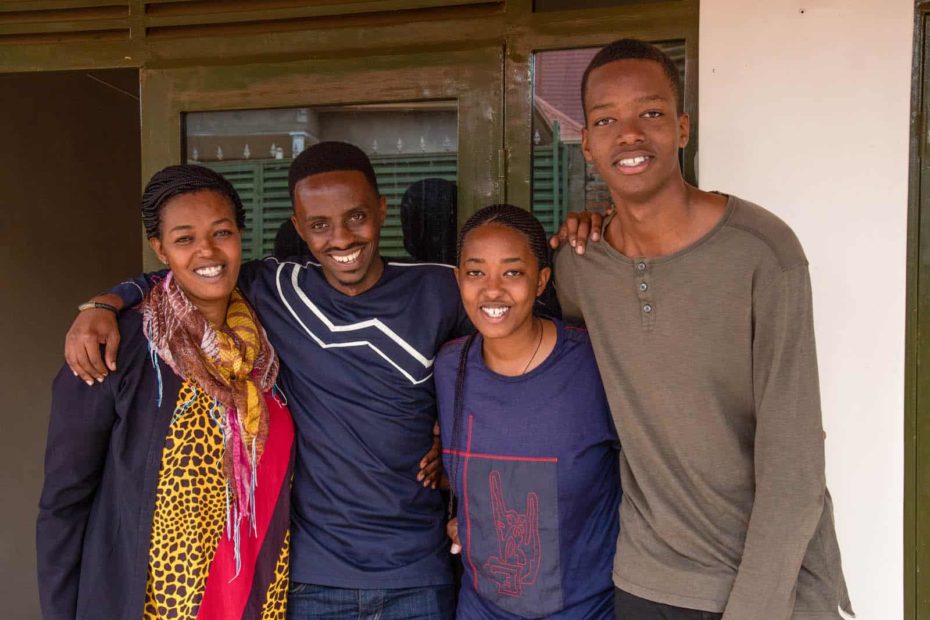 Christian is wearing a navy blue shirt with gray stripes down the center. He is outside his aunt's home with his Aunt Pascasie, and his cousins, Cynthia and Mugisha. They have their arms around each other.