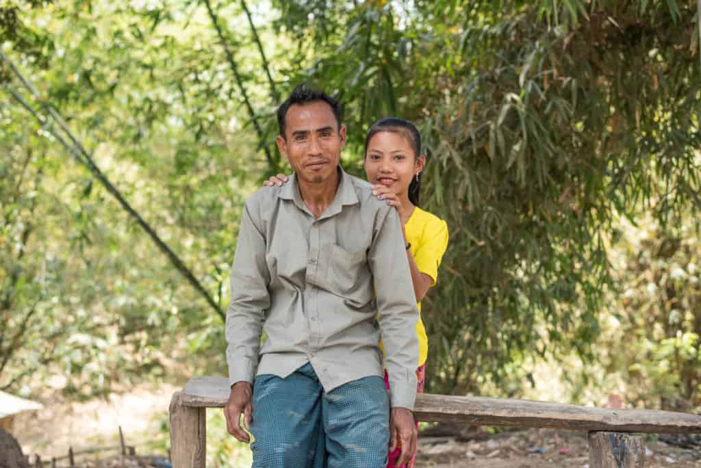 Baby is wearing a yellow shirt and is standing behind her father, who is wearing a tan shirt and blue pants. They are outside and there are bamboo trees in the background.