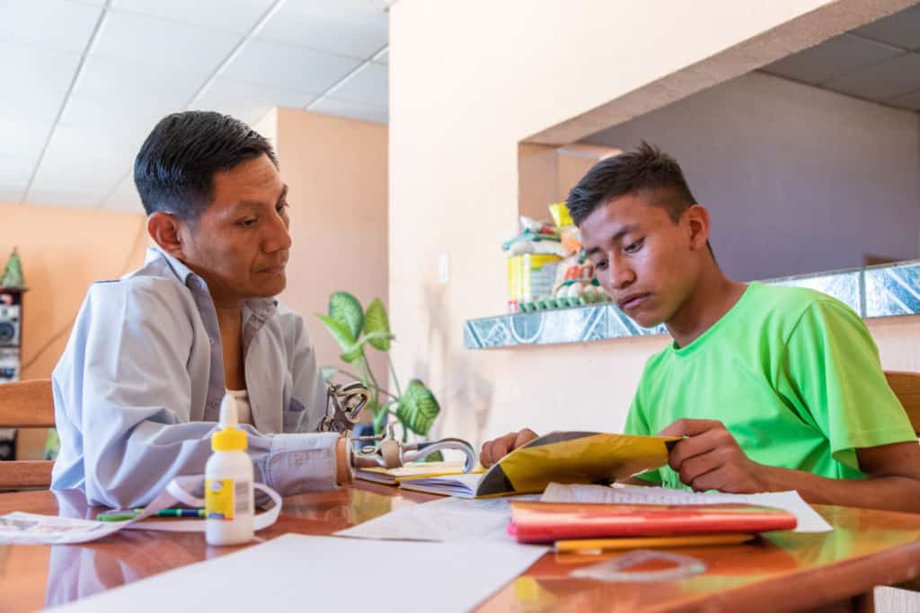 Jose is wearing a bright green shirt and black shorts. His father, Rene, is wearing a light blue shirt and jeans. They are sitting together at a table and Rene is helping Jose with his homework.