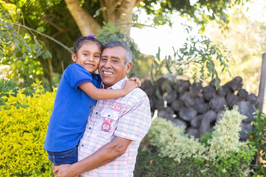Thatiana is wearing jeans and a dark blue shirt. Her father, Remberto, is wearing a plaid shirt and gray pants. Remberto is holding Thatiana. They are standing outside and there are trees in the background.