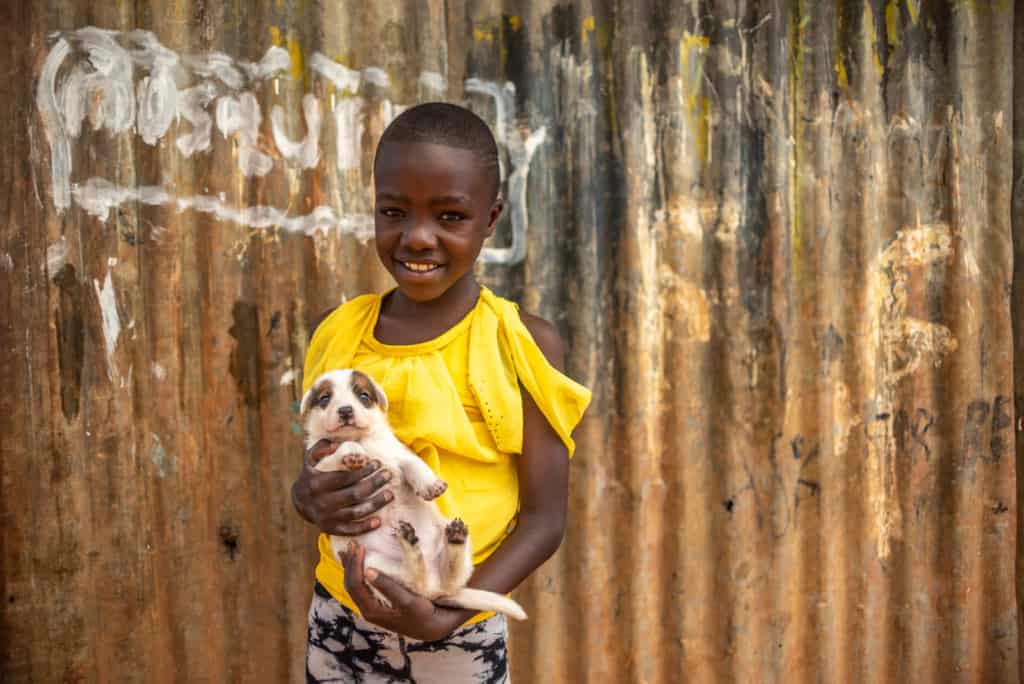 Leach is seen here wearing a bright yellow shirt, standing in front of a corrugated metal fence. She is holding a puppy, and is smiling at the camera.