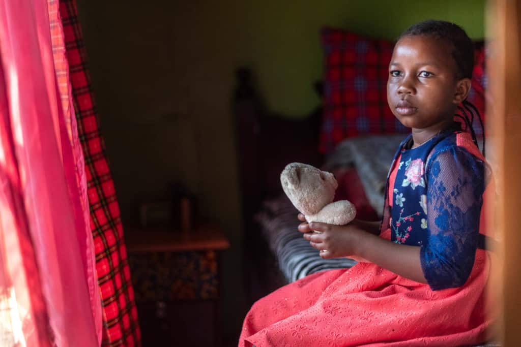 Shaniz is one of Kenya's girls. She is wearing a red dress with a blue floral print jacket. She is sitting on a bed in her home and is holding a teddy bear. There are red curtains in the window.