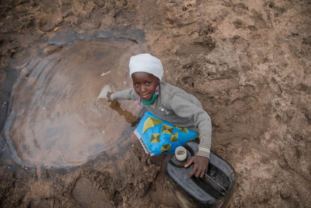 Faith is wearing a blue dress and a gray sweatshirt. They are bothShe is kneeling down and getting water from a dug out in the river.