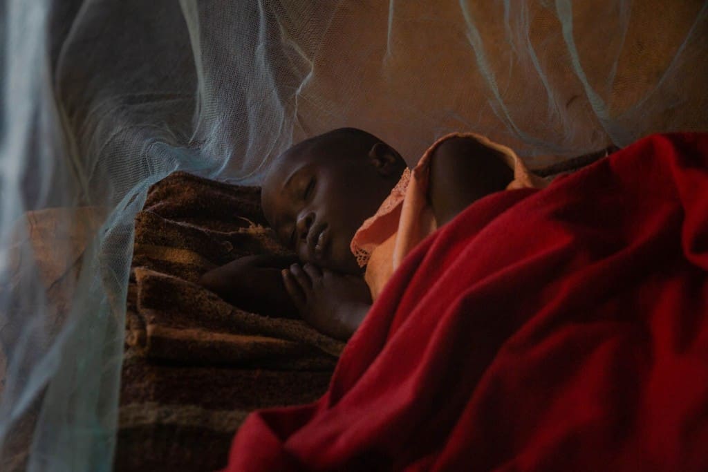 Abigail is sleeping on a bed under her mosquito net. There is a red blanket covering her.