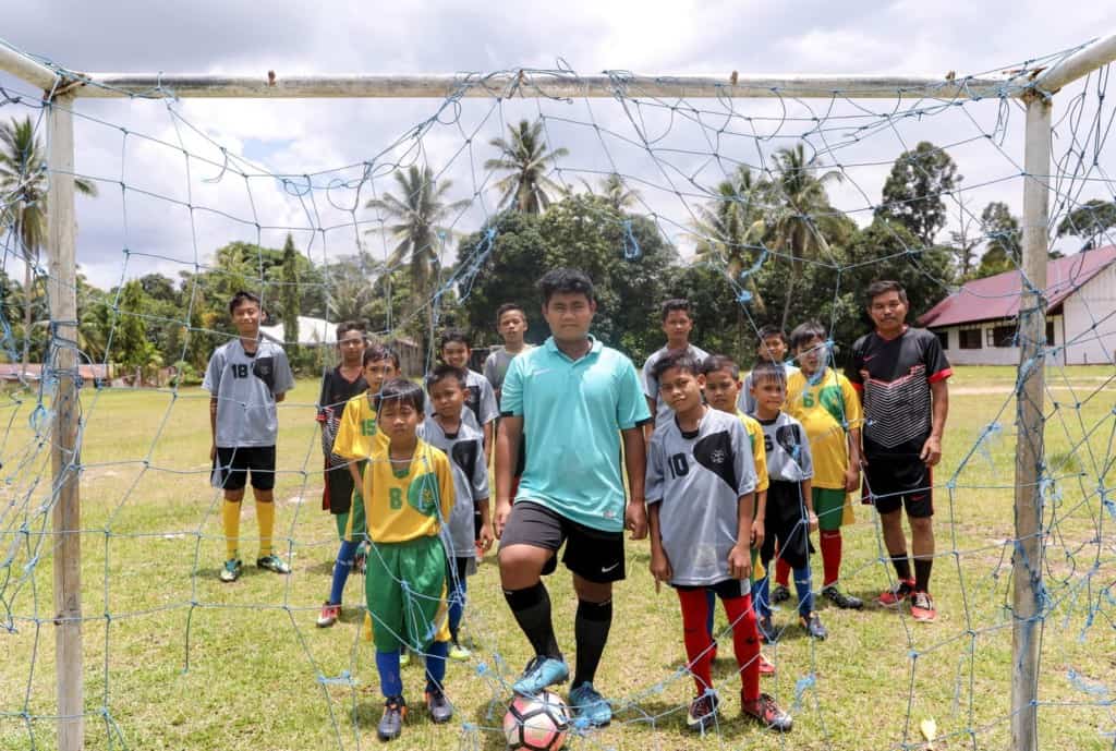  A group of boys standing together behind a ripped soccer goal, net. One is wearing mint t-shirt. Another is wearing black t-shirt on the right side.