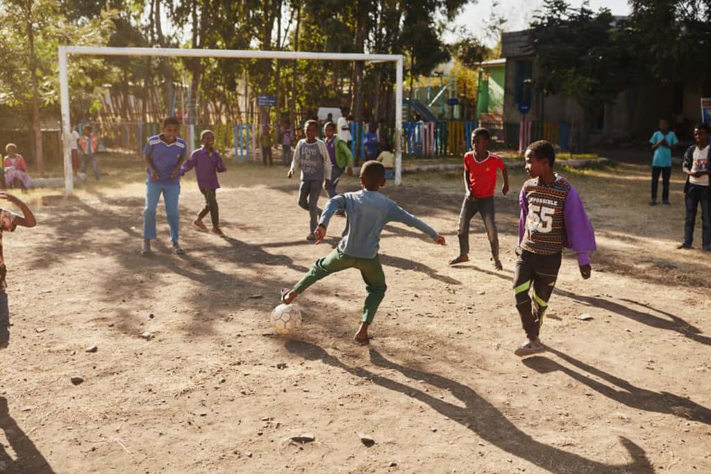 A group of boys are playing soccer on the dirt field outside the center. A boy in a blue shirt and green pants has his foot near the ball. There are trees and a white soccer goal in the background.