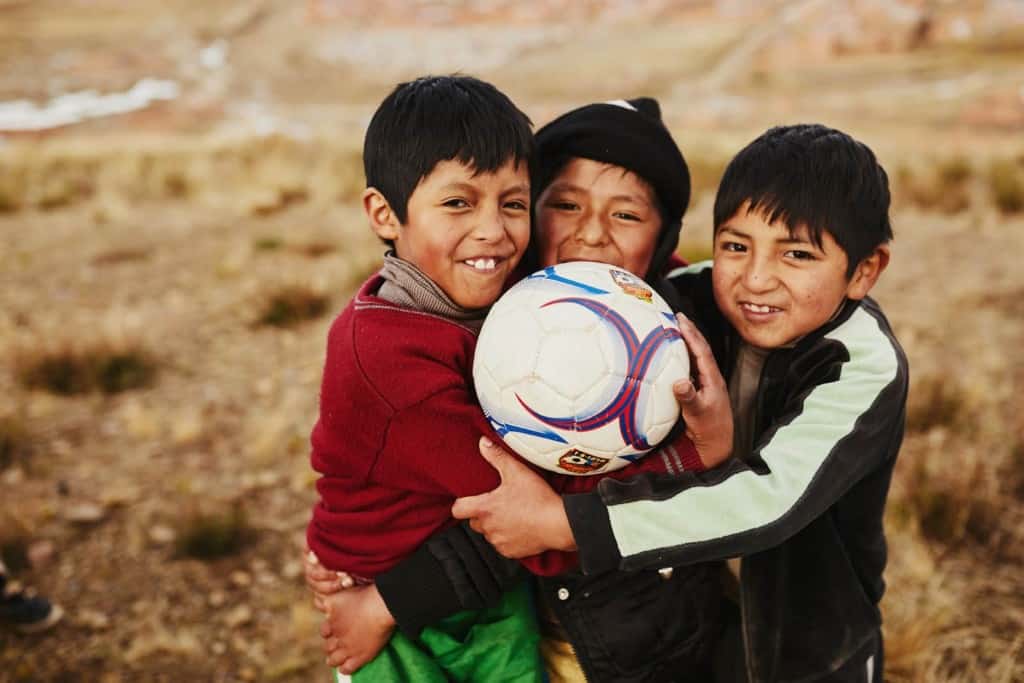 Three boys stand together on a dry grass field with their arms around each other holding a white soccer ball.