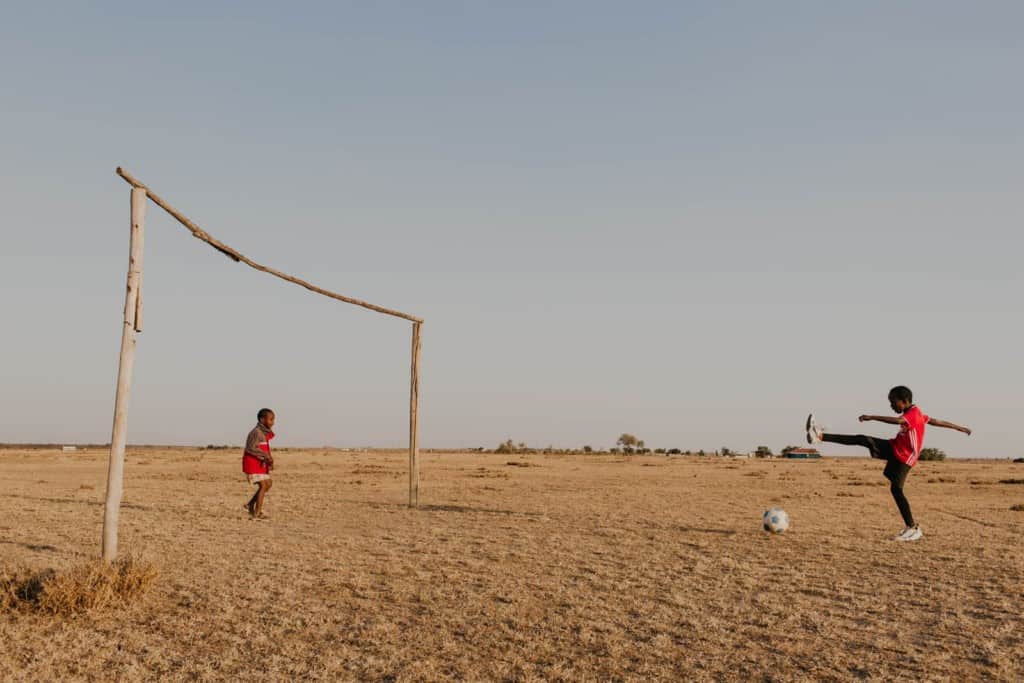 Praise, a 10 year old girl, wearing a red athletic sport shirt, kicks at a soccer ball on the dirt ground playing with another child.