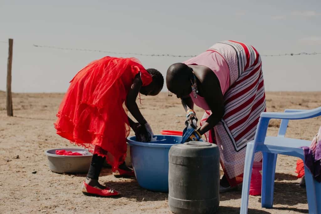 Praise, a 10 year old child, girl, sponsored child, wearing a red dress, bends over to help her mother, parent, adult woman, wash and clean dishes, daily household chores, outside using a large blue tub and soap. 