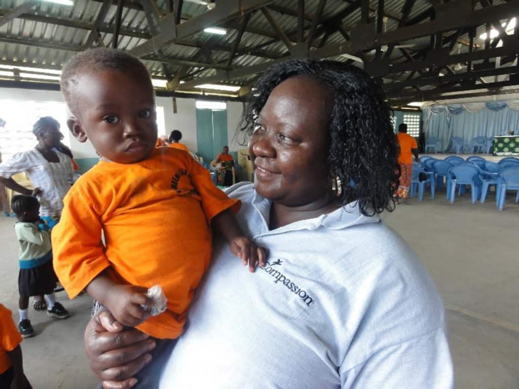 Susan, wearing a white shirt, is holding a toddler wearing a bright orange shirt.