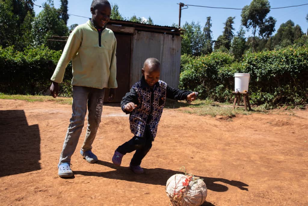 Young boy wearing a black shirt with a geometric pattern on the front and black pants. He is outside his home playing soccer with his older brother who is wearing jeans and a light green shirt.