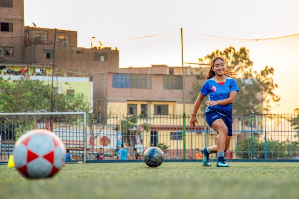 A 15-year-old, adolescent female, teenager, is practicing soccer on a soccer field. She is wearing a blue soccer uniform. Buildings and soccer goals are in the background.