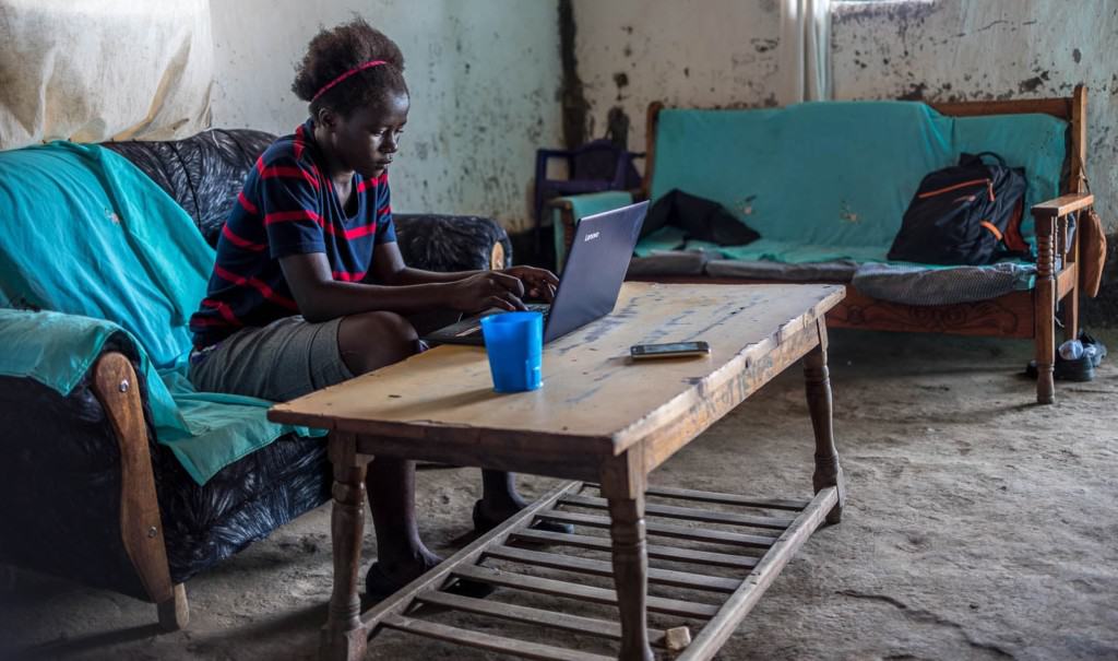 An adolescent girl in a navy blue and red striped shirt sits on a blue sofa at a coffee table looking at and using a black laptop computer with her hands on the keyboard. There are a blue cup and a cell phone on the table. There is another couch with a black backpack on it. The walls are white with black splatters on them.