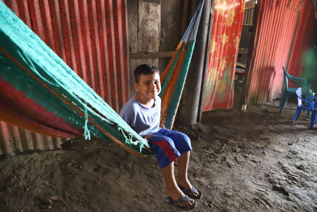 Boy sitting in a hammock. The background is the divisions inside the house, and behind them are the bedrooms where the family sleeps.