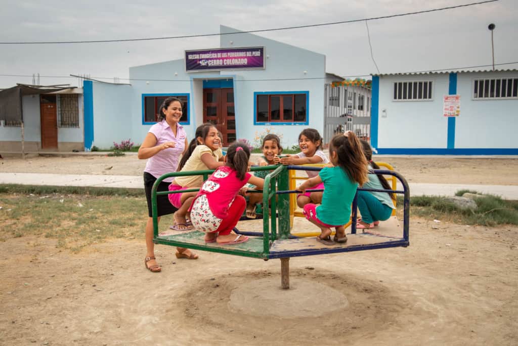 A group of children are playing outside on playground equipment with their tutor Iris Violeta, who is in a lavender shirt. The church building in the background is blue.