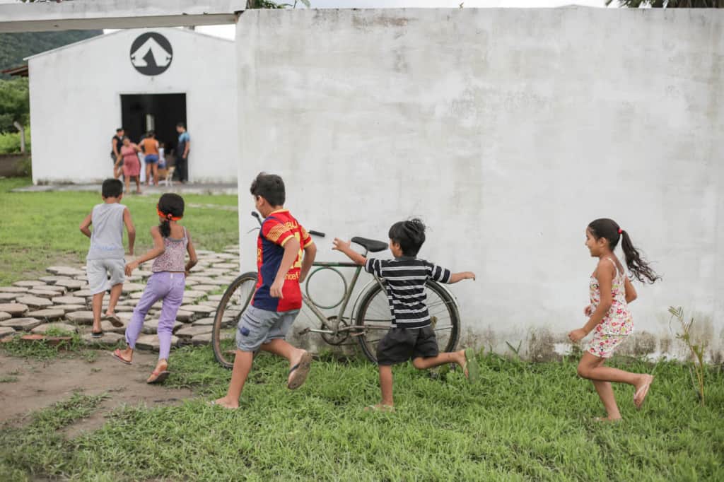 A group of children are running through the grass into the child development center, which is in a white building.