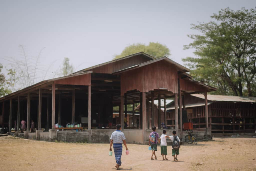Oosamai is walking behind his children Gungmae-ou (purple shirt) and his twin sons to drop them off at the project center, a large wood structure, building.