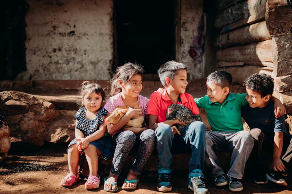 Amanda, in a blue and white shirt, Yessica, in pink, Argelio, in a bright orange-pink shirt, a boy in green, and a boy in blue are sitting on the steps in front of a building looking at each other and holding chickens. There are more chickens on the ground in front of them. The building is made of wood, corrugated metal an concrete.