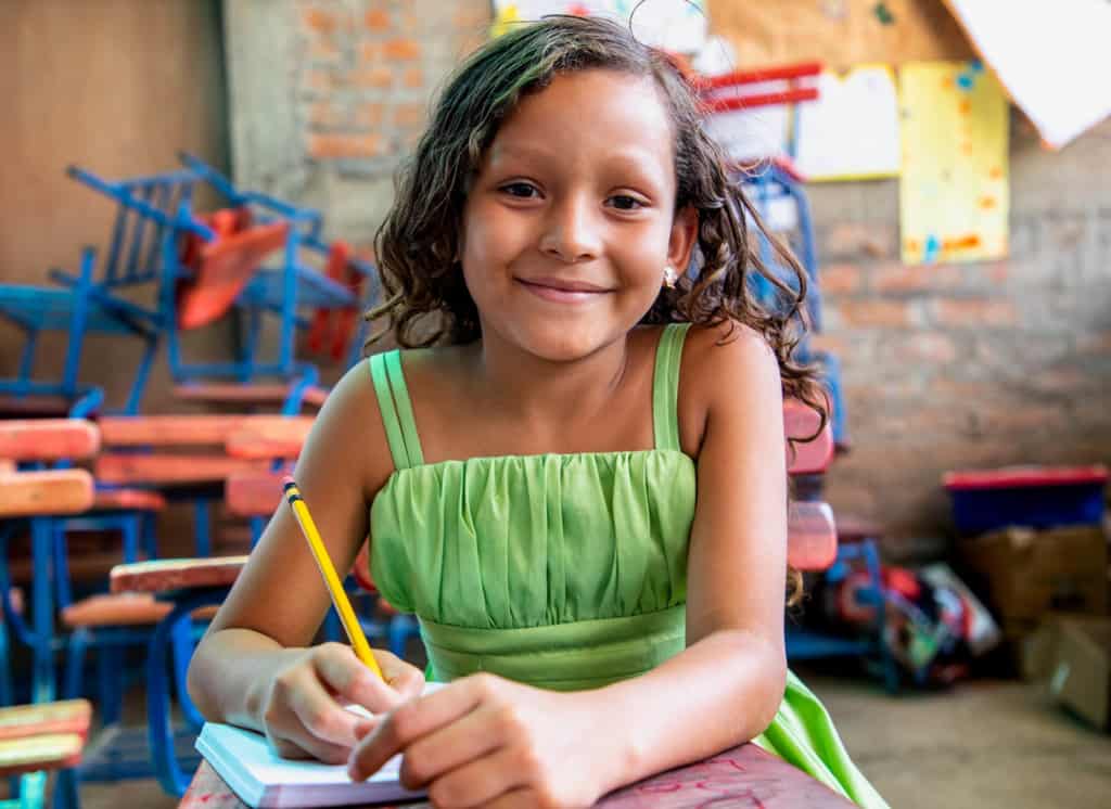 Young girl wearing a green dress and sitting at one of the desks inside one of the newly constructed classrooms. She is holding a pencil in her hand, and there is a notebook on her desk. In the background there are more desks and posters that the children made and hung from the walls. 