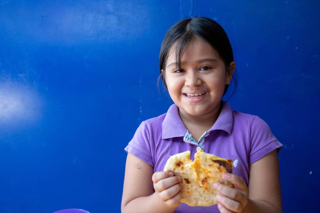 A 9 year old girl holds in her hand one ‘pupusa’ stuffed with cheese and shredded carrot. She smiles joyfully. She is wearing a purple polo shirt and has her hair in a ponytail. The background is a blue wall.