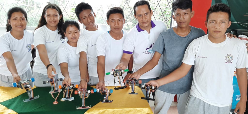 A group of young adults, all wearing white t-shirts, standing together showing their robotics projects.