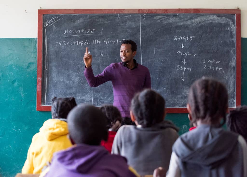 Daniel, wearing a purple sweater, is with a group of children at the Compassion project. The children are sitting at wooden desks and Daniel is standing in the front of the classroom. There is a chalkboard behind him.